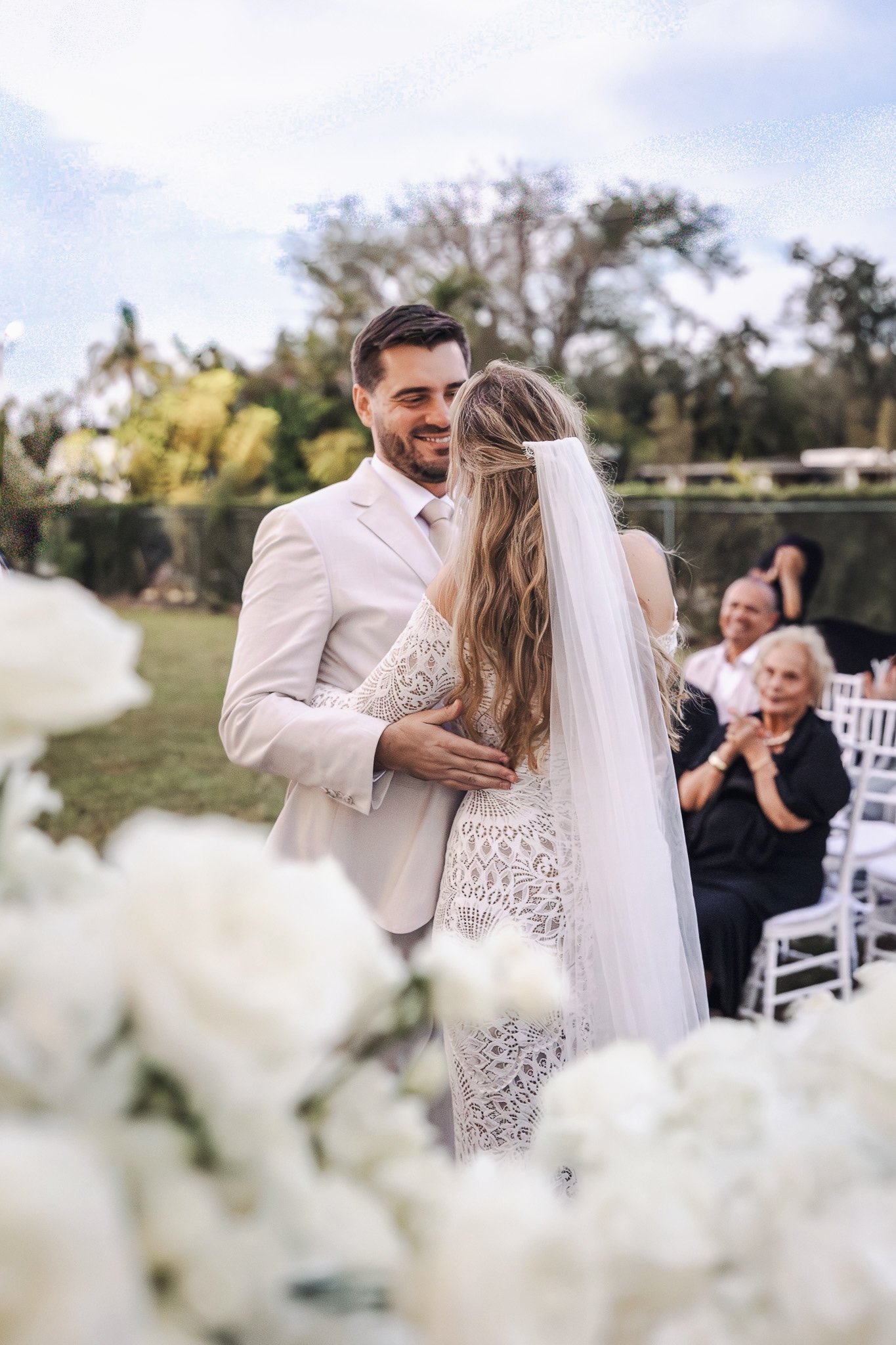 Bride and groom embracing during an outdoor ceremony framed by white florals